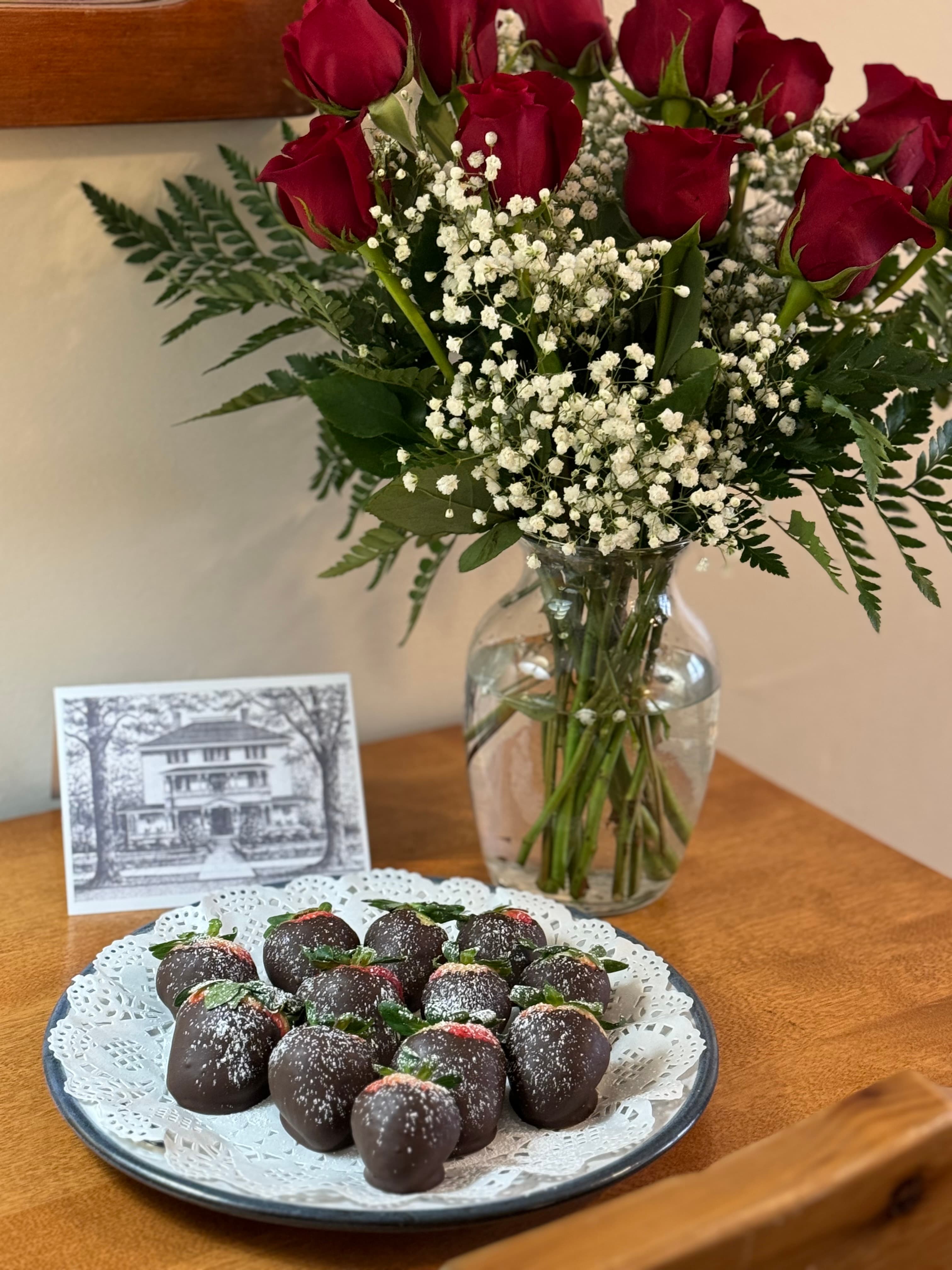 A plate of chocolate-covered strawberries is displayed beside a bouquet of red roses and a vintage photo in a glass vase on a wooden table.