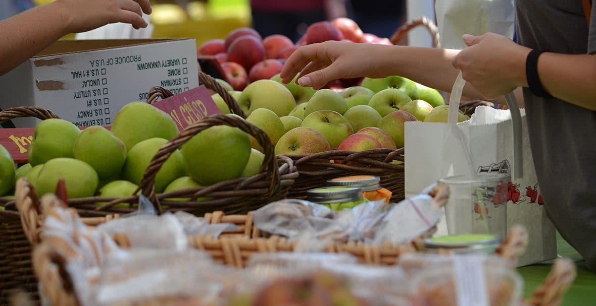 Hands reach for fresh green apples in a market setting with baskets of fruit and baked goods.