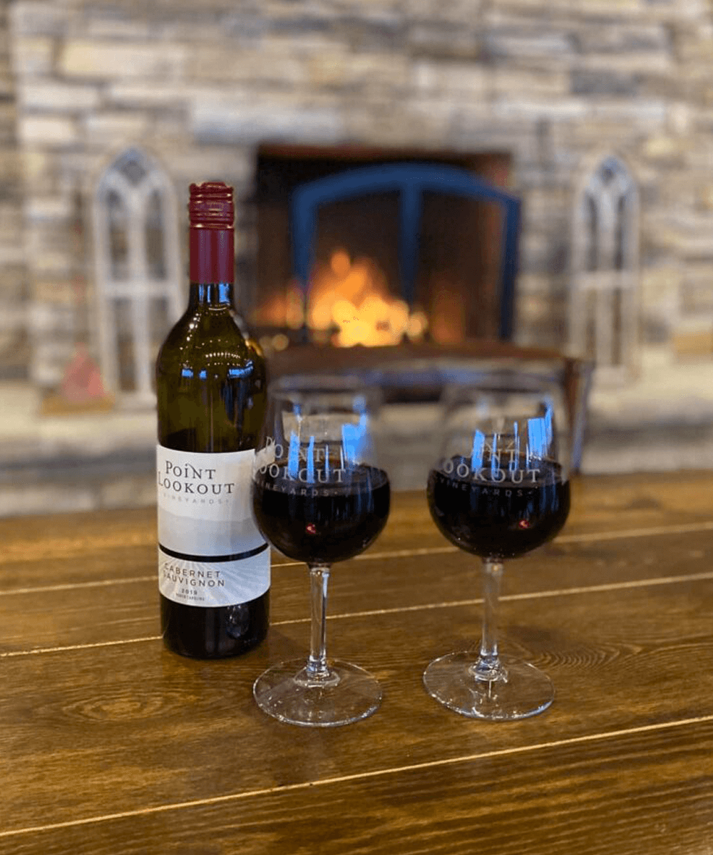 A bottle of Point Lookout Cabernet Sauvignon next to two filled wine glasses, set against a cozy stone fireplace.