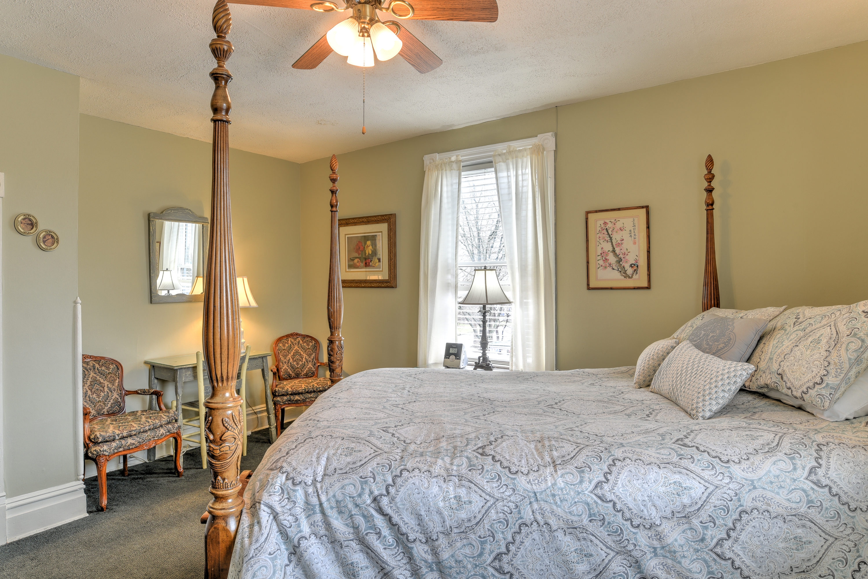 Room features queen four posted bed, and sitting area with two cushioned chairs and painted desk.