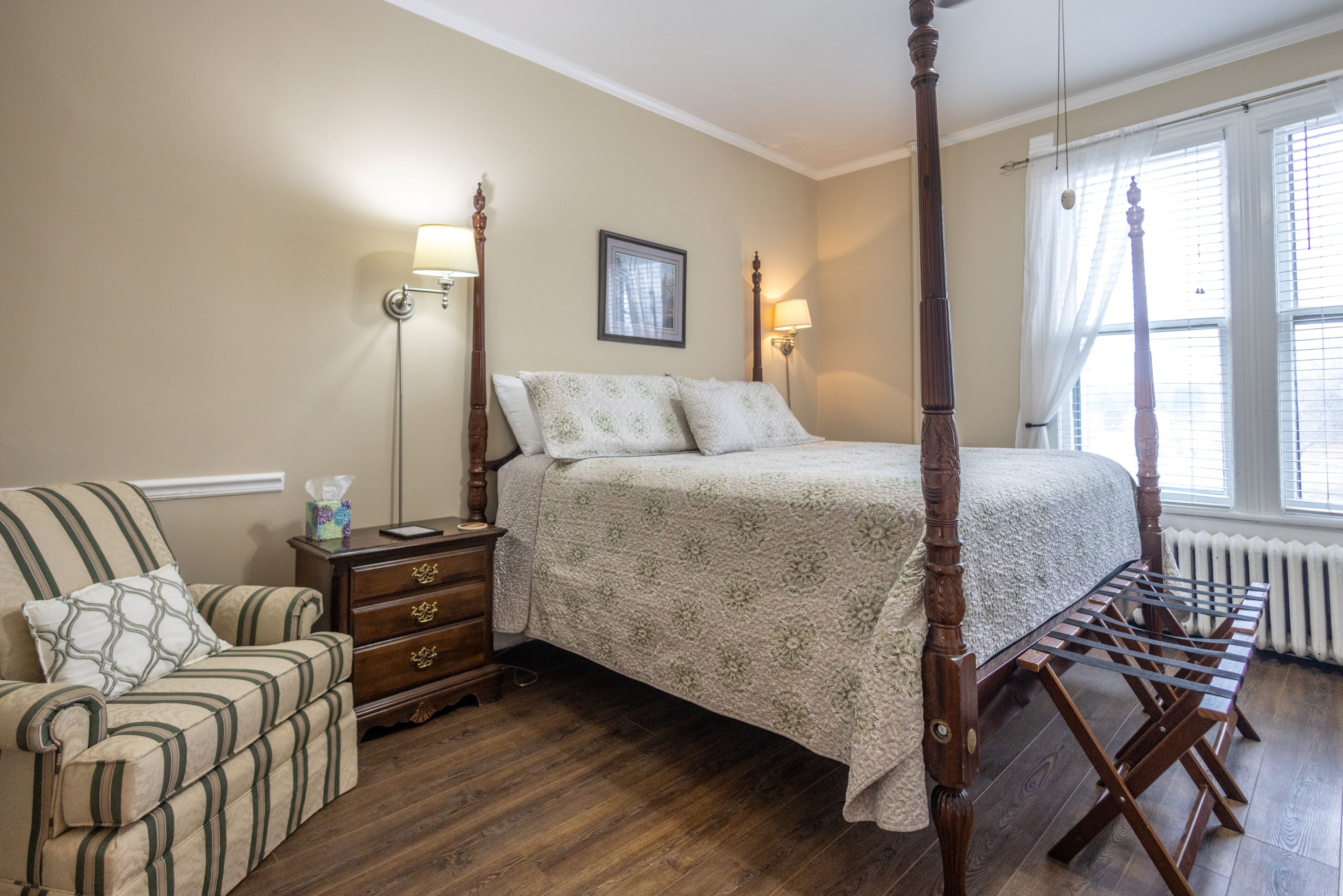 Room features wood flooring, light colored walls, a four posted king bed, and a striped comfy chair.