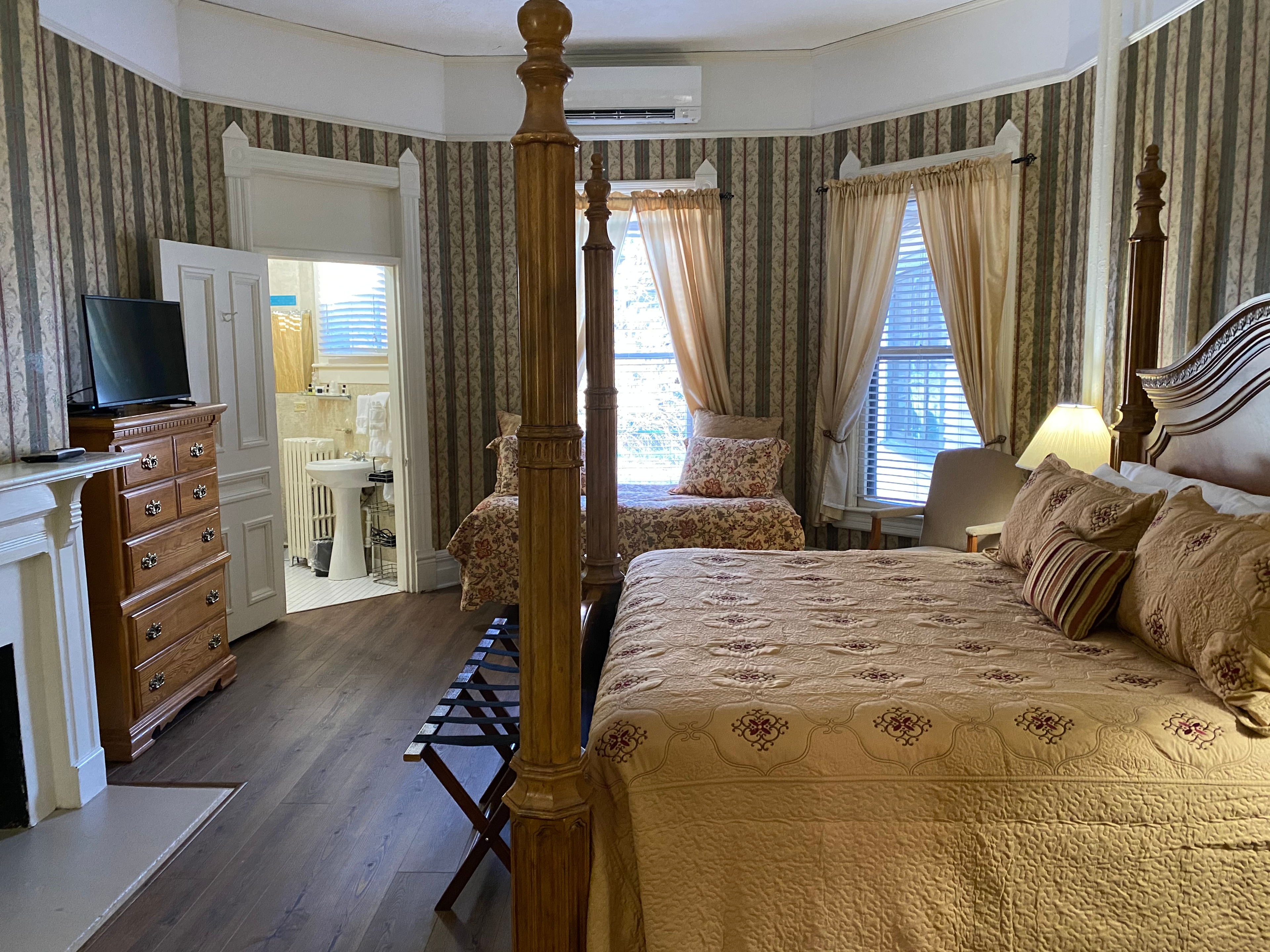 Room features wood flooring, striped wallpaper, a four posted king bed, twin bed, and tall wooden dresser with TV.