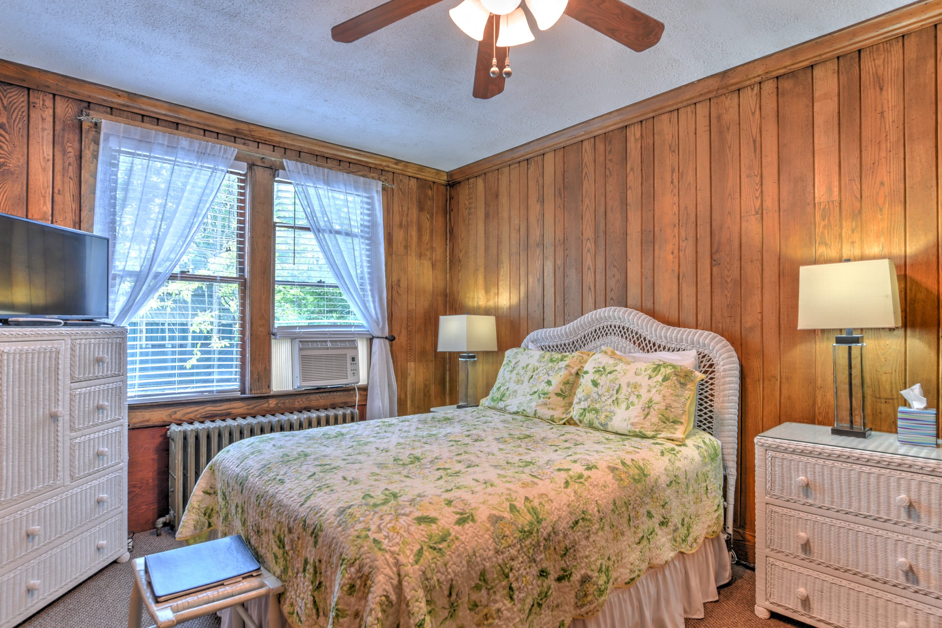 Room features Wormy Chestnut wood paneled walls and white wicker furniture, including night stands, dressers, and a queen bed.