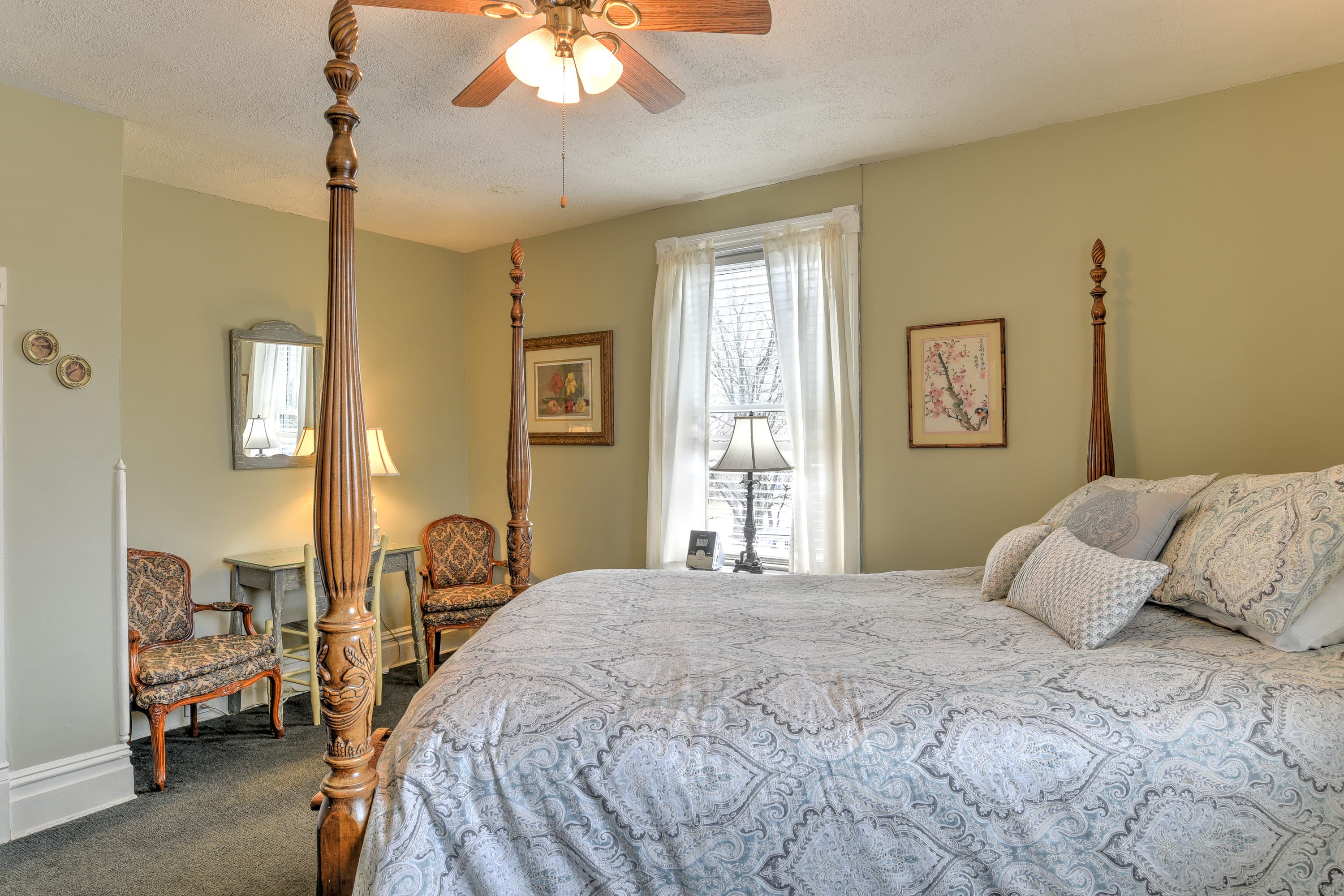 Room features queen four posted bed, and sitting area with two cushioned chairs and painted desk.