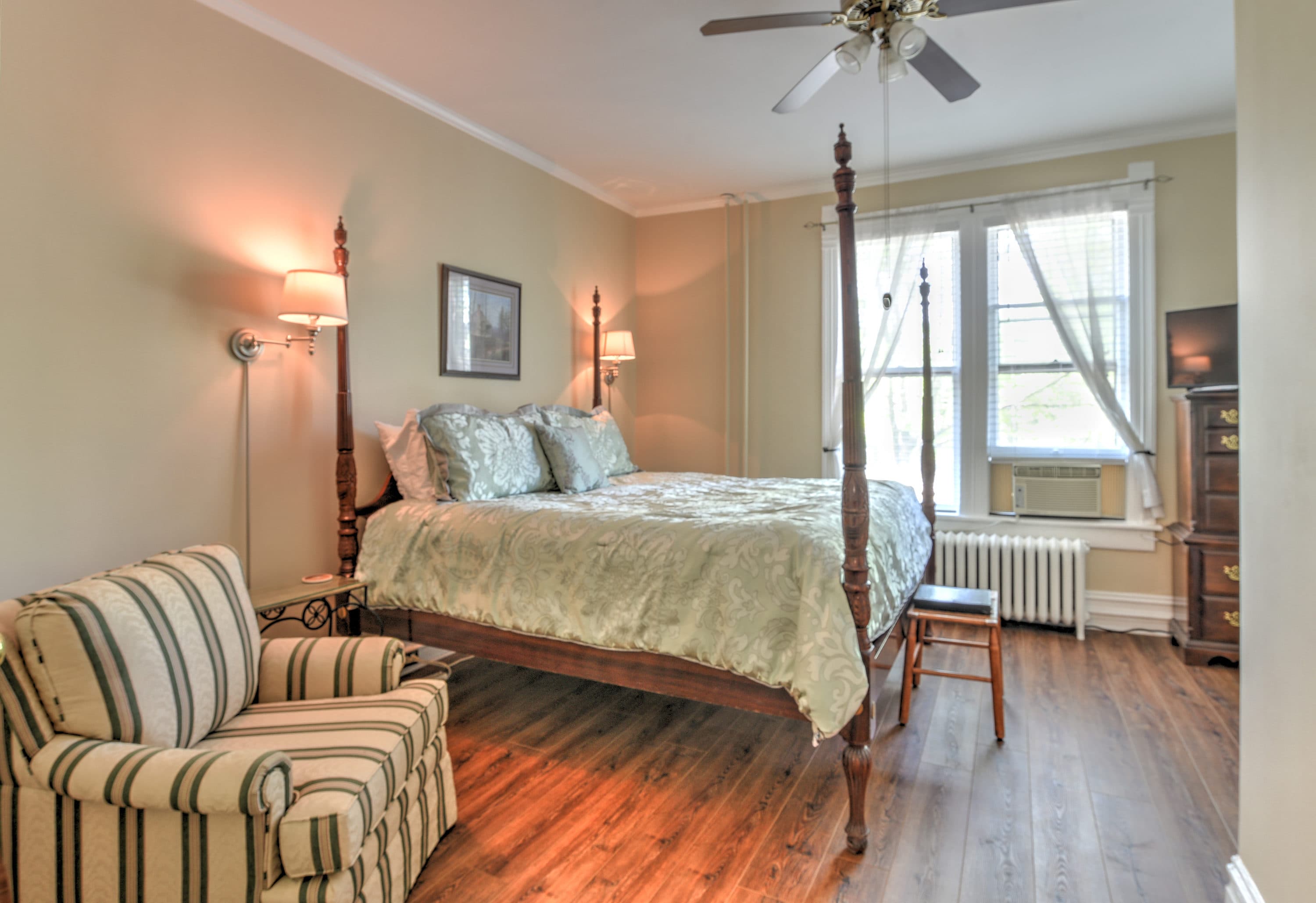 Room features wood flooring, light colored walls, a four posted king bed, and a striped comfy chair.