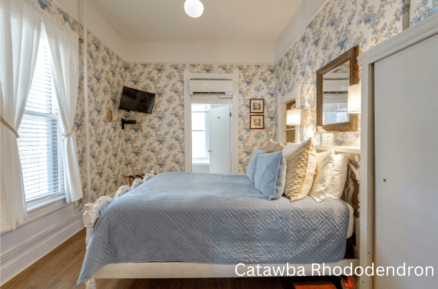 A cozy bedroom with floral wallpaper, a blue quilted bedspread, and a small TV mounted on the wall.