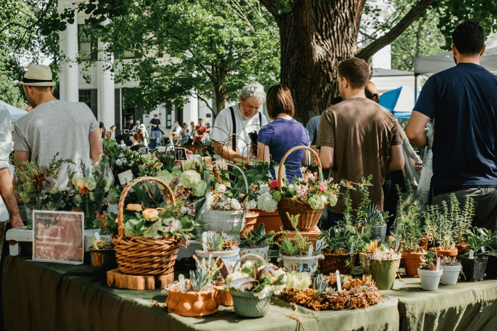 A busy outdoor market stall displays various flower and plant arrangements, with shoppers browsing.