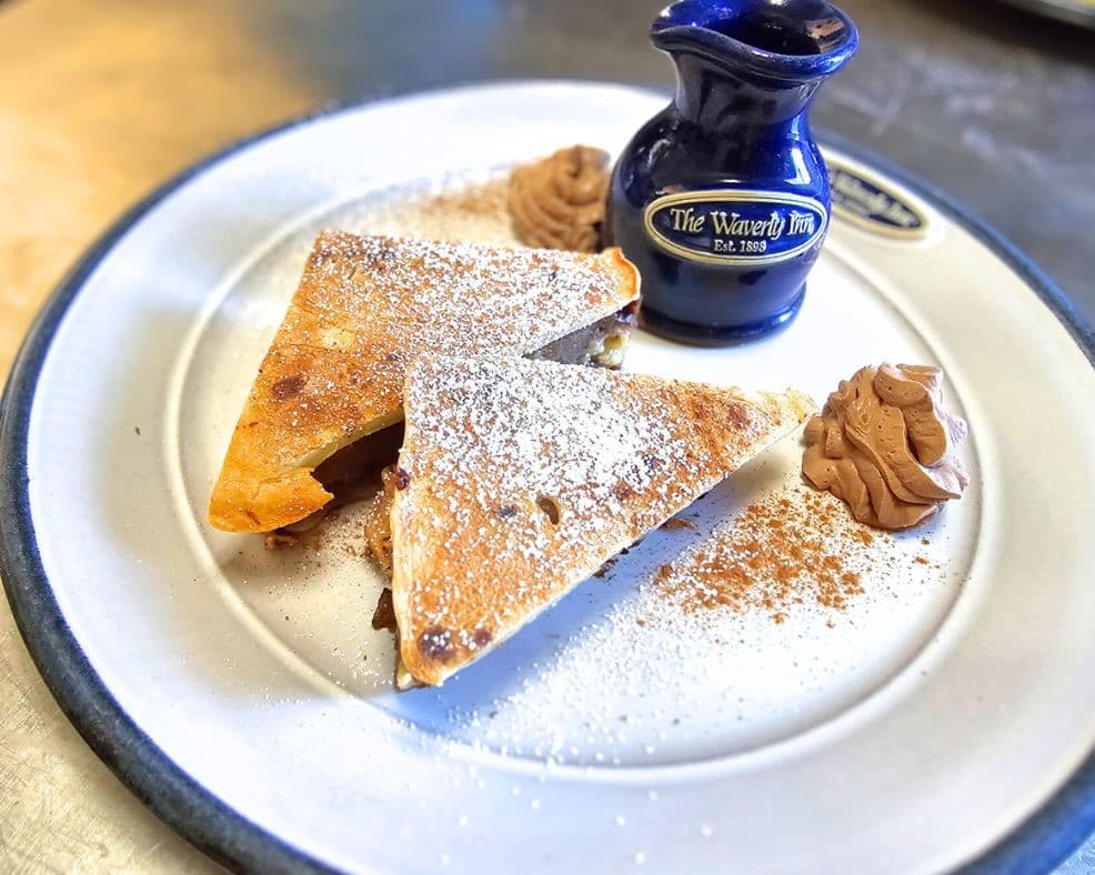 A plate featuring two triangular pieces of dessert dusted with powdered sugar, chocolate sauce, and whipped cream on the side.