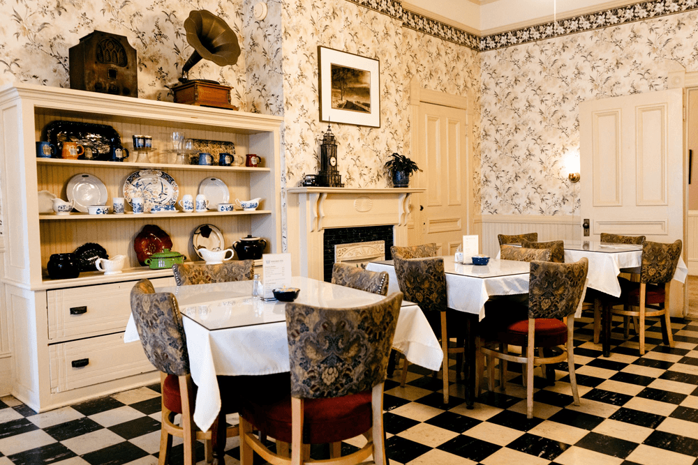 A cozy dining area with floral wallpaper, patterned chairs, and shelves displaying vintage dishware.