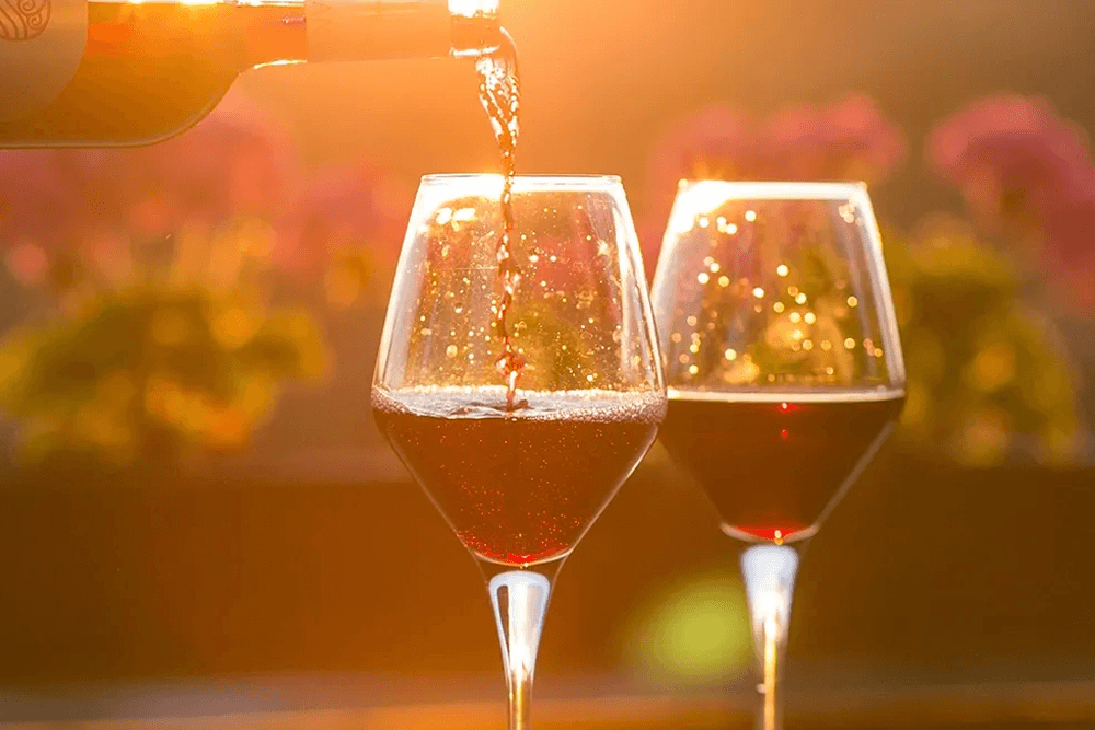 Red wine is being poured into two glasses against a sunlit background.